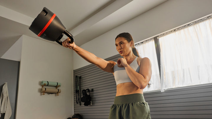 A woman holds a BowFlex SelectTech 840 Kettlebell in front of her while exercising in a home gym setting.