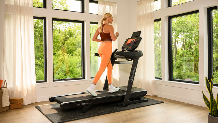 A woman in athletic wear runs on a BowFlex T16 Treadmill in a bright room with large windows and green outdoor views.