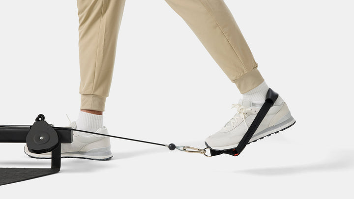Legs and feet, wearing beige joggers, white socks, and white sneakers, demonstrating the cable pulley system on the BowFlex Xtreme 2 SE Home Gym against a white background.
