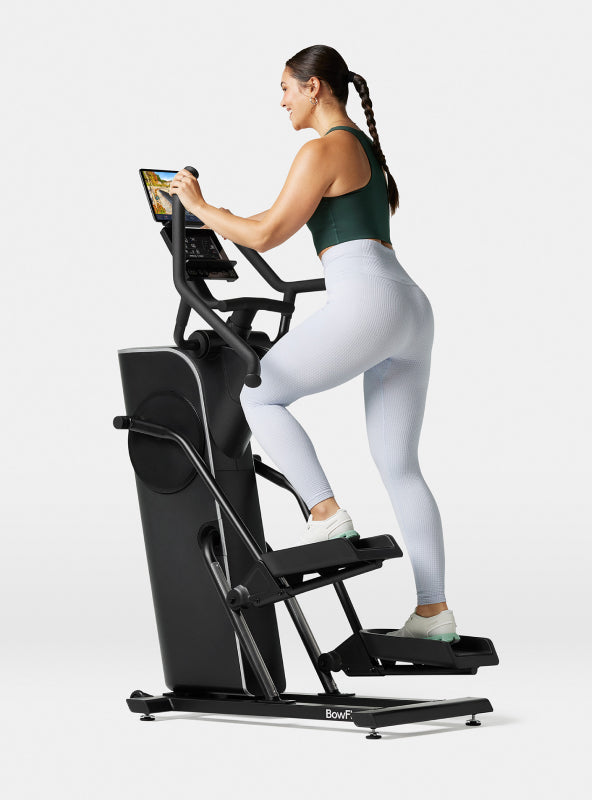 Angled side view of a woman exercising on the BowFlex Max Trainer SEi against a white background with a tablet on the product media shelf.