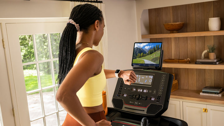 A woman in a home setting runs on a BowFlex T9 Treadmill with a tablet on its media holder showing a JRNY app screen with mountains.